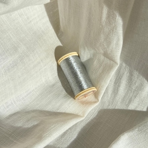 Könnte beinhalten: Eine Spule mit metallisch silbernem Garn und hellen Kunststoffenden. Der Faden ist fest um den zylindrischen Kern gewickelt, was eine reflektierende, schimmernde Oberflächenstruktur erzeugt. Die Spule liegt auf einem cremeweißen Stoffhintergrund.