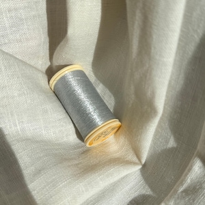 Könnte beinhalten: Eine Spule mit metallisch silbernem Garn und hellgelben Kunststoffenden. Der Faden ist fest um den zylindrischen Kern gewickelt, was eine reflektierende, schimmernde Oberflächenstruktur erzeugt. Die Spule ist schräg positioniert und zeigt den metallischen Glanz der silbernen Faser vor dem umgebenden Stoff.