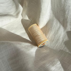 Pode incluir: Um carretel de linha de costura na cor creme com um núcleo cilíndrico de plástico e uma pequena etiqueta circular na extremidade. A linha está enrolada firmemente ao redor do carretel, exibindo um tom bege claro consistente.