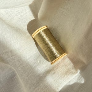 Könnte beinhalten: Eine Spule mit metallisch goldenem Garn mit glänzender, reflektierender Oberfläche, die um einen hellbeigen Kunststoffkern gewickelt ist. Die Spule liegt schräg und wirft einen deutlichen Schatten auf den umgebenden, strukturierten cremeweißen Stoff.