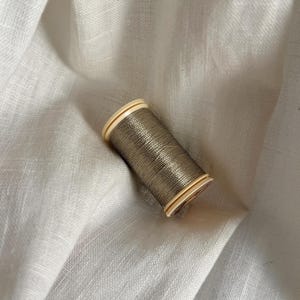 Könnte beinhalten: Eine Garnrolle aus metallisch olivgrünem Faden mit hellbeigen Kunststoffenden. Der Faden ist fest um den zylindrischen Kern gewickelt und weist einen subtilen Glanz sowie eine glatte, gleichmäßige Textur über die gesamte sichtbare Länge auf.