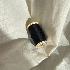 Könnte beinhalten: Eine schwarze Nähgarnrolle in zylindrischer Form mit hellen Kunststoffenden. Das obere Ende ist mit einem runden Etikett versehen, das eine kleine Illustration und Text zeigt, wobei der dunkle Faden fest um den Kern gewickelt ist.