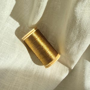 Könnte beinhalten: Eine Spule mit metallischem Goldfaden, der eine glänzende, reflektierende Oberfläche aufweist und um einen hellbeigen Kunststoffkern gewickelt ist. Der Faden ist fest in zylindrischer Form aufgewickelt und zeigt eine lebendige, warme Goldfarbe.