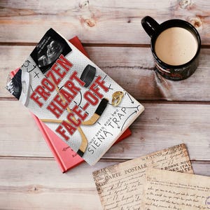 May include: A book titled "Frozen Heart Face-Off" with a hockey stick and a puck on the cover. The book is sitting on a pink surface with a postcard and a coffee mug on a wooden surface.
