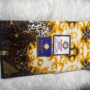 May include: A white fabric with a gold and black baroque pattern and a leopard print. The fabric has a blue label with the text "ROCK TEX CREATION PRINTED BY ROCK TEX" and a gold label with the text "PREMIUM QUALITY ROCK PRINTED BY ROCK TEX".
