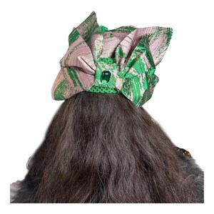 May include: A decorative head covering with a green and pink abstract pattern. The head covering has a bow shape with a green jewel in the centre. The head covering is on top of dark brown hair.