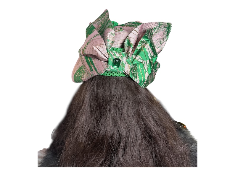 May include: A decorative head covering with a green and pink abstract pattern. The head covering has a bow shape with a green jewel in the centre. The head covering is on top of dark brown hair.