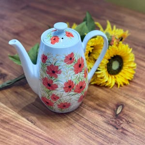 May include: A light blue ceramic teapot with a lid, decorated with red poppy flower designs. The teapot has a curved spout and handle. A bouquet of yellow sunflowers rests behind the teapot on a wooden surface.