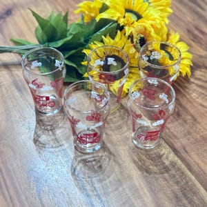 May include: Five clear glass tumblers featuring a red vintage car design and white clouds. The glasses are set on a wooden surface with a bouquet of yellow sunflowers in the background. The tumblers are a vintage style.