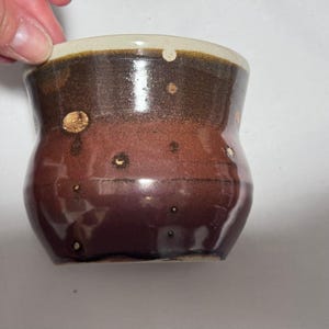 Peut inclure: Une tasse en céramique faite à la main, de forme organique unique. La tasse présente un dégradé de couleurs, passant d'une base marron foncé à un milieu brun-rouge, et un bord beige clair. De petits accents mouchetés ajoutent de l'intérêt visuel.