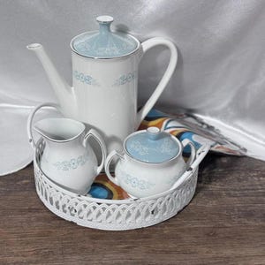 May include: A white porcelain tea set with a light blue floral design, including a teapot, creamer, and sugar bowl, presented on a white, ornate, oval tray. The set is placed on a wooden surface with a colorful patterned cloth.