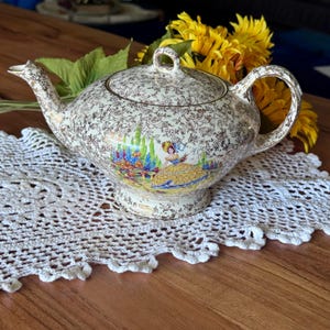 Peut inclure: Théière vintage crème avec un motif floral et une scène peinte d'une femme en robe jaune. La théière a un bec verseur incurvé, un corps arrondi et un couvercle assorti. La poignée est arquée et décorée du même motif floral.