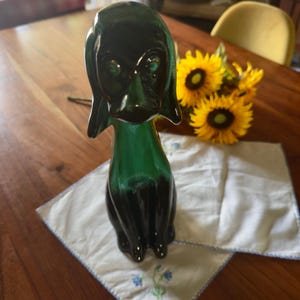 May include: A dark green glass figurine shaped like a dog with long, drooping ears and a smooth, reflective surface. The dog is depicted in a seated position with a slender neck and a rounded head, showcasing a deep emerald hue that transitions to black.