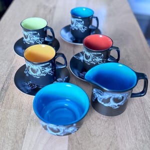 Peut inclure: Un ensemble de tasses et soucoupes en céramique noire avec un motif floral. L'intérieur des tasses est de différentes couleurs, dont le jaune, le vert, le rouge et le bleu. Un pot à crème et un bol assortis sont également inclus.