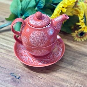 May include: A coral-coloured ceramic teapot and teacup set with a matching saucer. The teapot and cup feature a white, intricate mandala pattern. The set is displayed on a wooden surface, with sunflowers and greenery in the background.