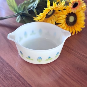 May include: A white, oval Pyrex bowl with two handles and a leaf pattern in blue and green. The bowl is on a wooden surface with yellow sunflowers. The bowl is likely vintage and suitable for kitchen use.