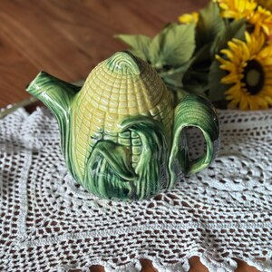 Peut inclure: Théière en céramique en forme d'épi de maïs, avec des détails jaunes et verts. La théière présente un motif détaillé de grains et de feuilles de maïs, avec une anse et un bec verseur. Posée sur une nappe en dentelle blanche, avec des tournesols en arrière-plan.