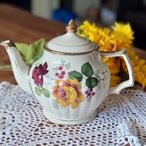 May include: A cream-coloured porcelain teapot with gold-coloured handle, spout, and trim. The teapot is decorated with a floral pattern featuring yellow and burgundy roses, green leaves, and small pink flowers. The lid has a gold finial.