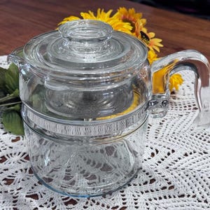 May include: A clear glass coffee pot with a lid and handle. The pot has a metal band around the middle and a filter basket inside. The handle is curved and the pot is sitting on a white lace doily.