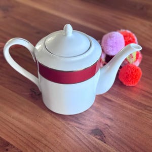 May include: White ceramic teapot with a red band and gold trim. The teapot has a curved handle and spout, and a lid with a small knob. A cluster of colourful pom-poms sits nearby. The teapot is on a wooden surface.