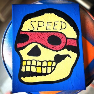 May include: A yellow skull with black outlines and a red bandana with white polka dots. The bandana has the word "SPEED" written in black across the top. The skull has gold teeth.