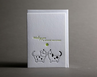 May include: White greeting card with the text "Wishing you a speedy recovery" in green script. The card features a die-cut design of two cartoon dogs, one wearing a cone. A green dot is above the dogs.