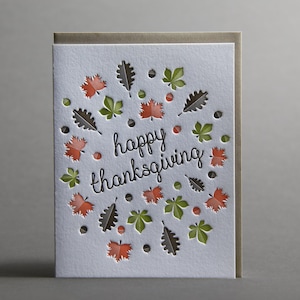 May include: A Thanksgiving greeting card with a white textured surface. The card features a design of cut-out leaves in orange, green, and brown, surrounding the words "happy thanksgiving" in black script. The card is set against a neutral background.