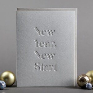 May include: A white greeting card with the embossed text "New Year, New Start". The card is surrounded by gold and blue decorative ornaments. The card has a textured surface.