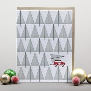 May include: A Christmas card featuring a pattern of grey and white Christmas trees. A red car carrying a Christmas tree is depicted on the card. The card is surrounded by Christmas baubles.