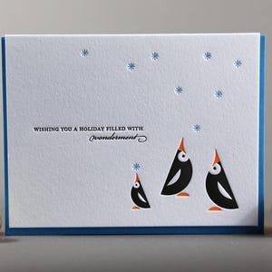 May include: A white holiday card with a blue border features three stylized black penguins with orange beaks and feet. The card has embossed snowflakes and the text "Wishing you a holiday filled with wonderment."