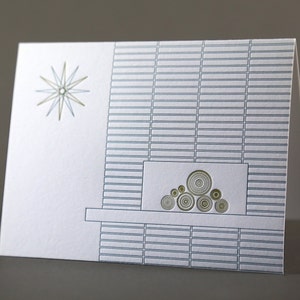 May include: A folded white greeting card with a mid-century modern design. The card features a starburst and a fireplace illustration with a brick pattern in shades of gray and gold. The card is blank inside.