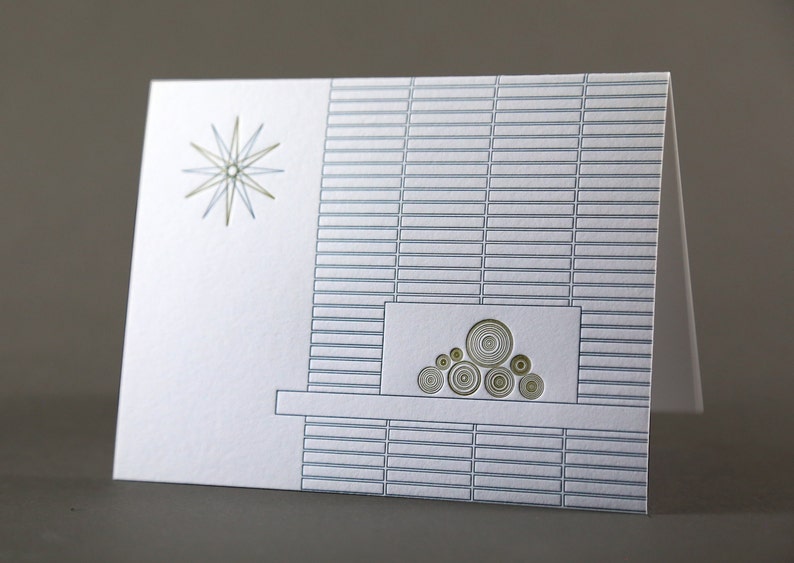 May include: A folded white greeting card with a mid-century modern design. The card features a starburst and a fireplace illustration with a brick pattern in shades of gray and gold. The card is blank inside.
