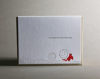 May include: A greeting card with a textured white surface and a beige border. The text "CONGRATS ON YOUR NEW DIGS!" is printed in black. Two circular designs and a red floral accent are on the bottom right.