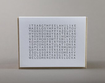 May include: A white rectangular greeting card featuring a grid of black letters arranged in a word search puzzle format. The text includes words such as thank you, happy, love, missing you, birthday, congratulations, anniversary, sorry, celebrate, welcome, and cheers.