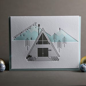 Könnte beinhalten: Eine weiße Grußkarte mit einem schwarz-hellblauen Berg- und Hüttendesign. Die A-Rahmen-Hütte hat einen Schornstein und ist von Bäumen umgeben. Die Karte ist mit goldenen und blauen Kugeln verziert.