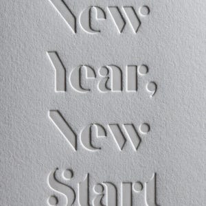 May include: A textured, off-white paper with the embossed words "New Year, New Start". The text is in a stylized, sans-serif font. The image is a close-up, highlighting the tactile quality of the paper and the typography.