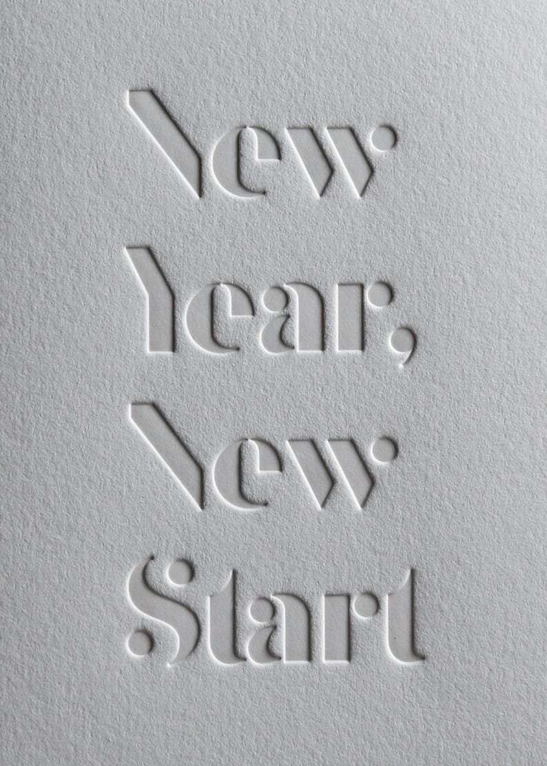 May include: A textured, off-white paper with the embossed words "New Year, New Start". The text is in a stylized, sans-serif font. The image is a close-up, highlighting the tactile quality of the paper and the typography.