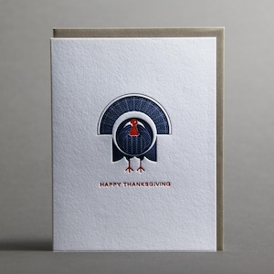 May include: Thanksgiving greeting card featuring a stylized turkey illustration in navy blue and red. The turkey has a fan-shaped tail and is centered on a textured white card. The words "HAPPY THANKSGIVING" are printed in red below the turkey.
