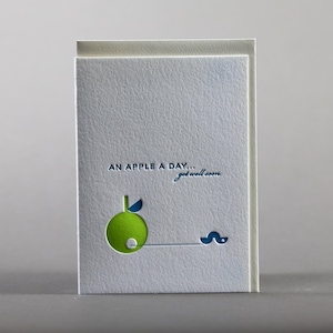 Könnte beinhalten: Eine Genesungskarte mit einer strukturierten, hellgrauen Hülle. Die Karte zeigt ein grünes Apfeldesign mit einem blauen Blatt und einem Wurm. Der Text "AN APPLE A DAY... get well soon." ist blau gedruckt.