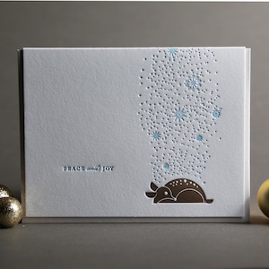May include: A white holiday card with the words "Peace and Joy" in a light blue font. The card features a brown deer illustration and a pattern of blue snowflakes and dots. Gold and glittery gold ornaments are on the left and right.