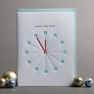 May include: A white New Year's greeting card with a clock design, red hands, and the words "Happy New Year!" in red. The card is surrounded by gold and silver decorative balls.