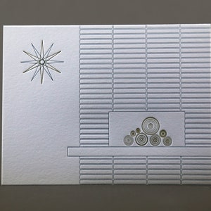 May include: A silver greeting card with a mid-century modern design. The card features a starburst and a fireplace motif with concentric circles. The background has a textured, rectangular pattern.