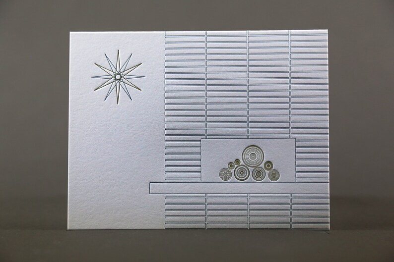 May include: A silver greeting card with a mid-century modern design. The card features a starburst and a fireplace motif with concentric circles. The background has a textured, rectangular pattern.