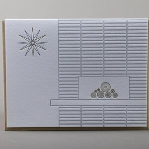May include: A greeting card with a minimalist design. The card features a starburst graphic on the left and a fireplace illustration on the right. The fireplace design includes a series of horizontal lines and circular shapes. The card has a neutral color palette.