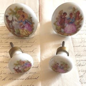 Peut inclure: Quatre boutons de placard ronds en porcelaine blanche ornés de scènes colorées représentant des personnages en tenue d'époque. Chaque bouton possède une base en métal doré et une vis de fixation, avec des illustrations détaillées de personnages.