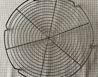 May include: Circular metal cooling rack with a concentric wire design. The rack is made of thin, dark-colored metal wires arranged in a radial pattern, with a series of curved edges. This kitchen tool is designed for cooling baked goods.