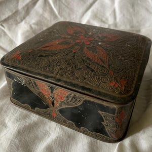 Puede incluir: Una caja de lata rectangular vintage, negra con tapa. La tapa presenta un diseño floral detallado en rojo y negro. Los lados de la caja también tienen detalles florales rojos. La caja tiene un aspecto ligeramente desgastado.