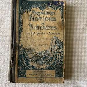 Può includere: Un libro antico intitolato "Premières Notions de Sciences" con una copertina usurata. La copertina presenta un'illustrazione di paesaggio in verde acqua e beige, con testo in francese. Il dorso del libro è scuro e le pagine mostrano segni di età.