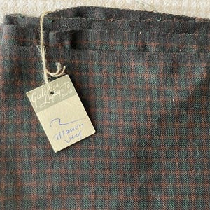 Könnte beinhalten: Nahaufnahme eines gefalteten Stoffes mit kleinem Karomuster in Braun-, Grün- und Grautönen. Ein Etikett von Galeries Lafayette Paris ist angebracht, mit dem handschriftlichen Text "Manoir".