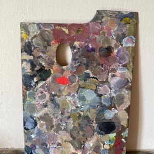 May include: A rectangular wooden artist palette featuring a thumb hole and a notched corner, covered in a dense, multi-layered accumulation of dried oil paint in various shades of grey, brown, blue, purple, and a single vibrant red spot.
